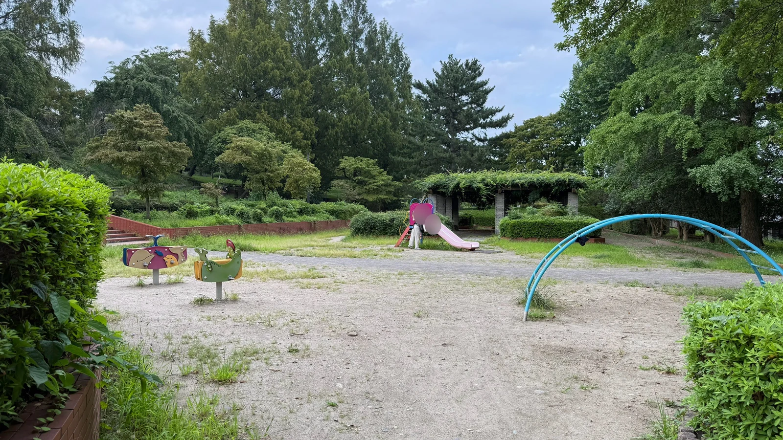 木々と生け垣に囲まれた、小さなすべり台や揺れる遊具、アーチ形の遊具がある広場