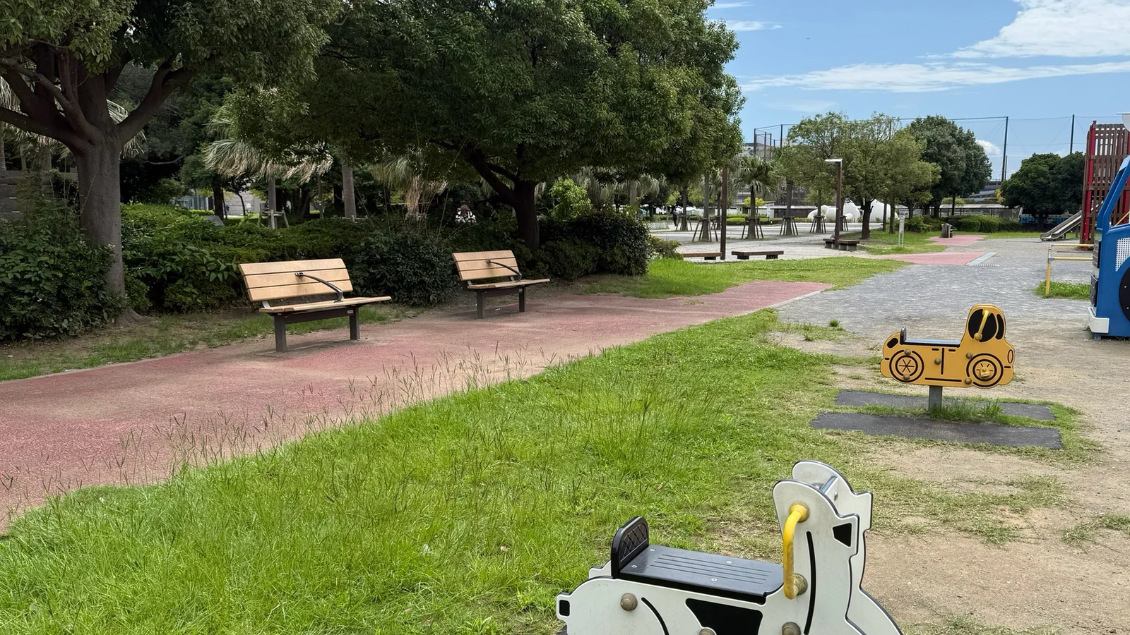 芝生に置かれたバイクの形の揺れる遊具と、奥のベンチや大型遊具